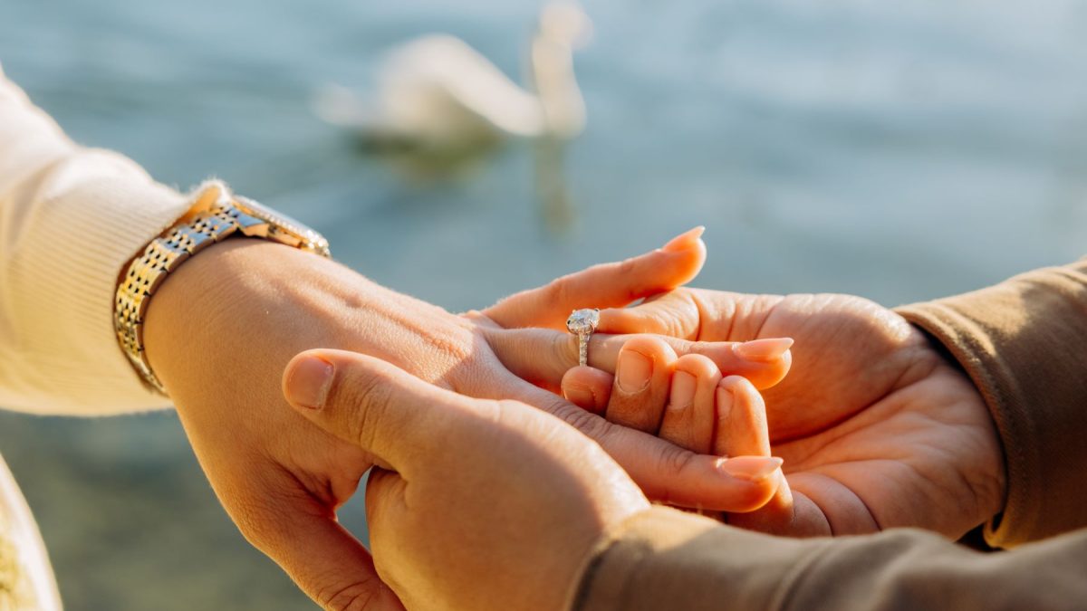 Hands exchanging a luxury engagement ring, with a soft-focus background of a serene outdoor setting.
