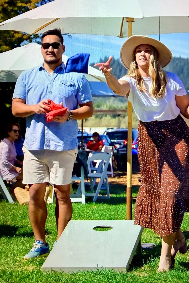 A woman throws a bean bag while a man stands nearby, both enjoying a game outdoors under umbrellas.