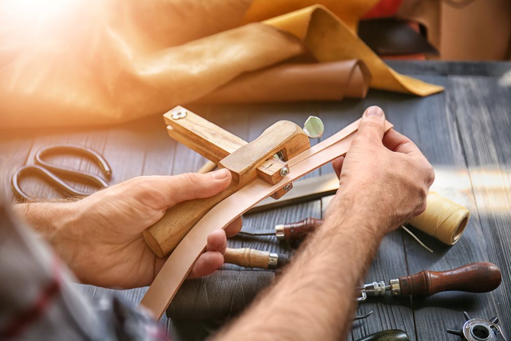 Hand adjusting a piece of leather with crafting tools on a wooden table, showcasing the process of belt making.