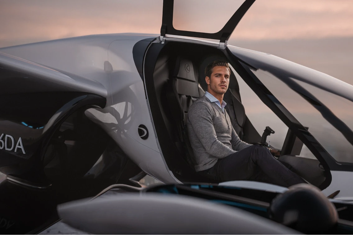 A man sits inside a futuristic aircraft with its canopy open, set against a sunset backdrop.