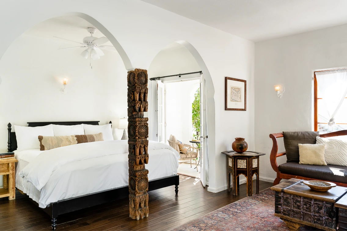 Bright and airy bedroom featuring a carved wooden pillar, white bedding, and a cozy seating area with natural light.