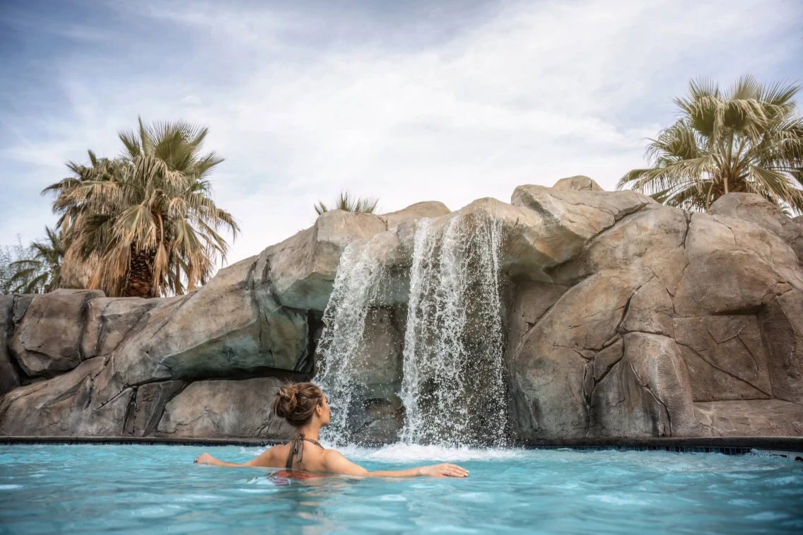Woman relaxing in a pool under a waterfall, surrounded by palm trees in a serene desert setting of Palm Springs.