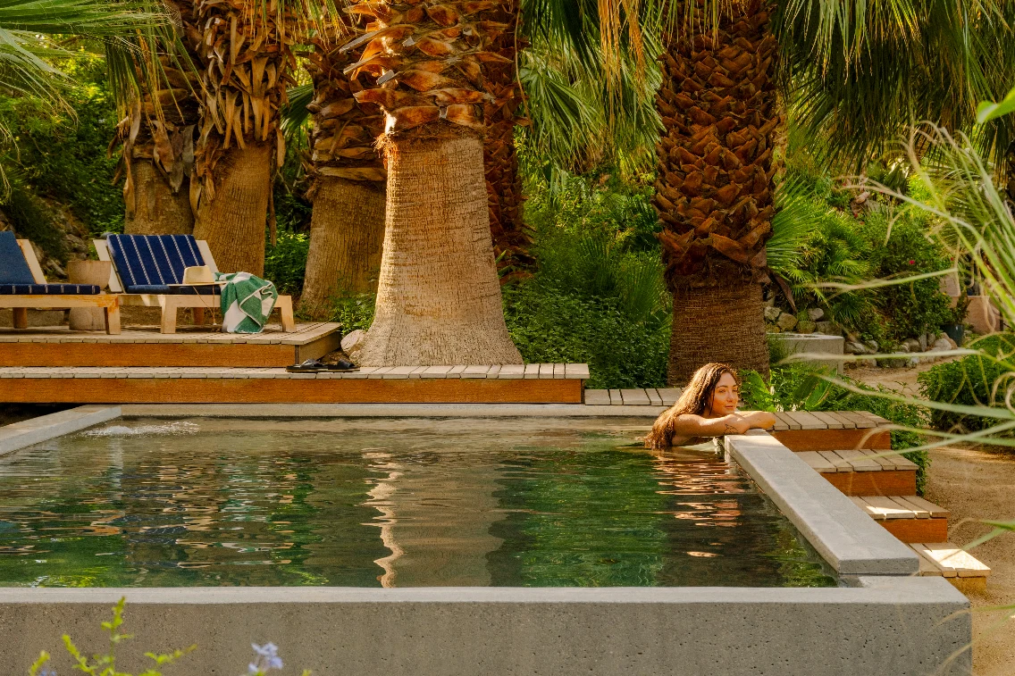 A person relaxes in a tranquil pool surrounded by palm trees and lounge chairs in a lush desert setting.