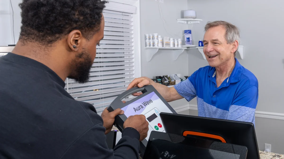 A customer receives the Aura Wave device from a staff member in a wellness center, discussing a noninvasive approach to health.