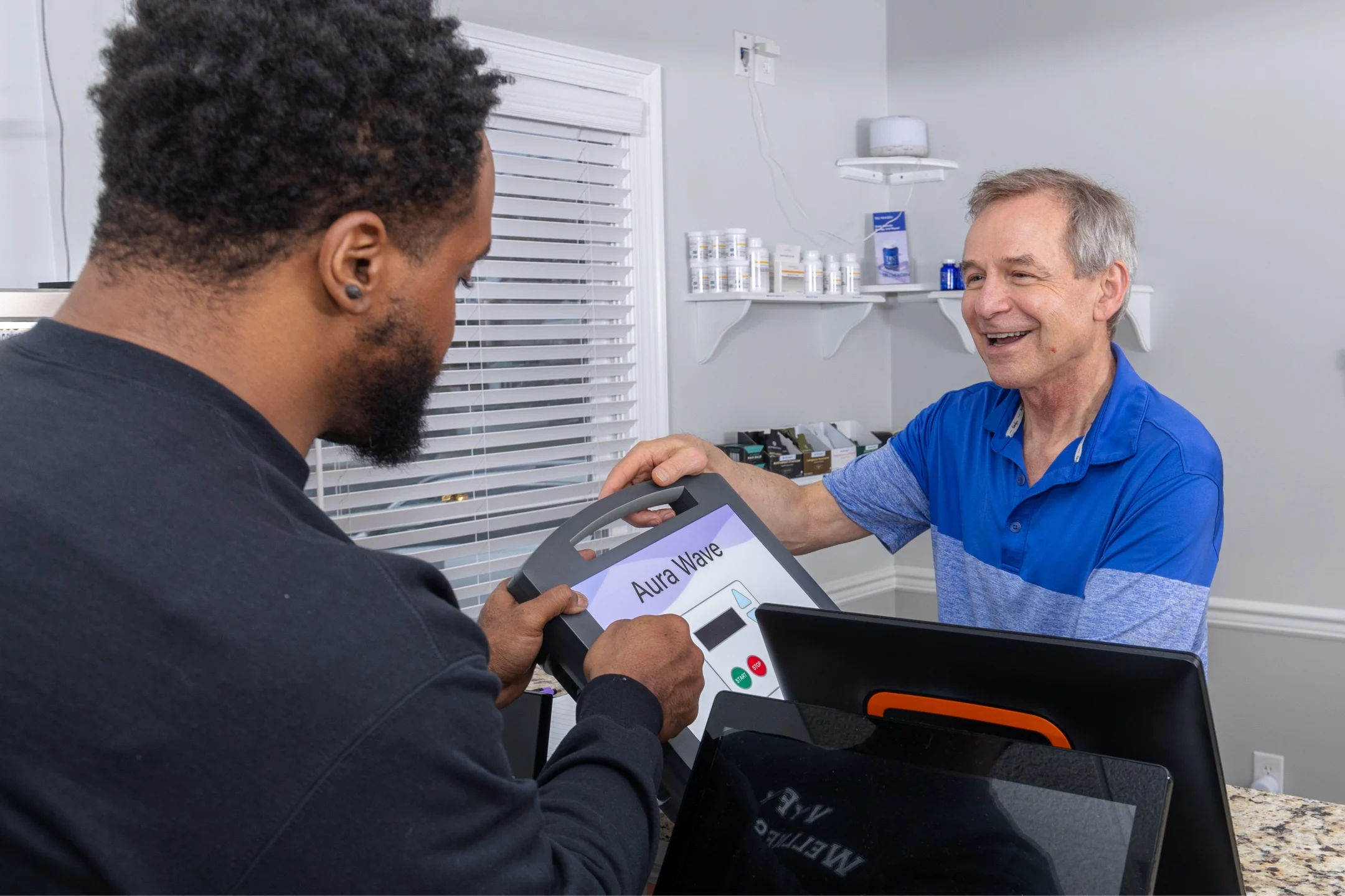 A customer receives the Aura Wave device from a staff member in a wellness center, discussing a noninvasive approach to health.
