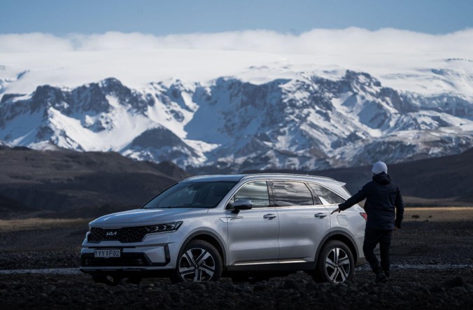 A person standing next to a silver SUV against a backdrop of snow-capped mountains in Iceland.