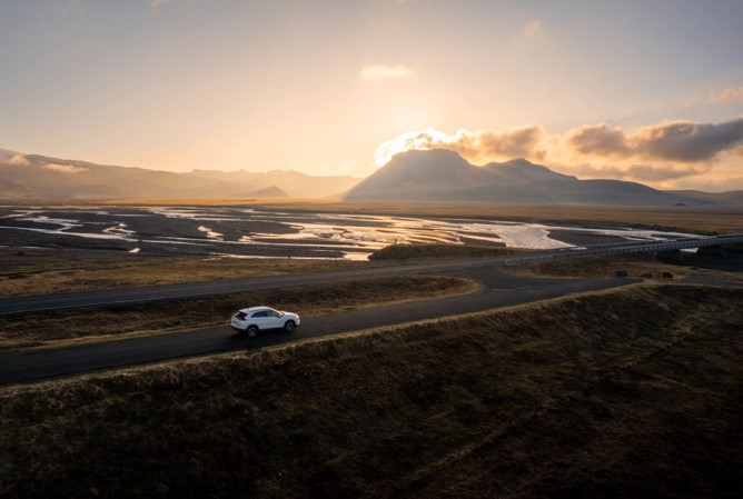 A car drives along a road in Iceland, surrounded by mountains and a winding river at sunset.