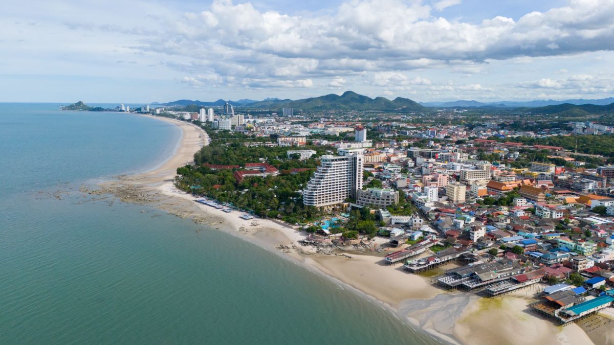 Aerial view of Hua Hin's coastline featuring sandy beaches, city buildings, and lush greenery under a partly cloudy sky.