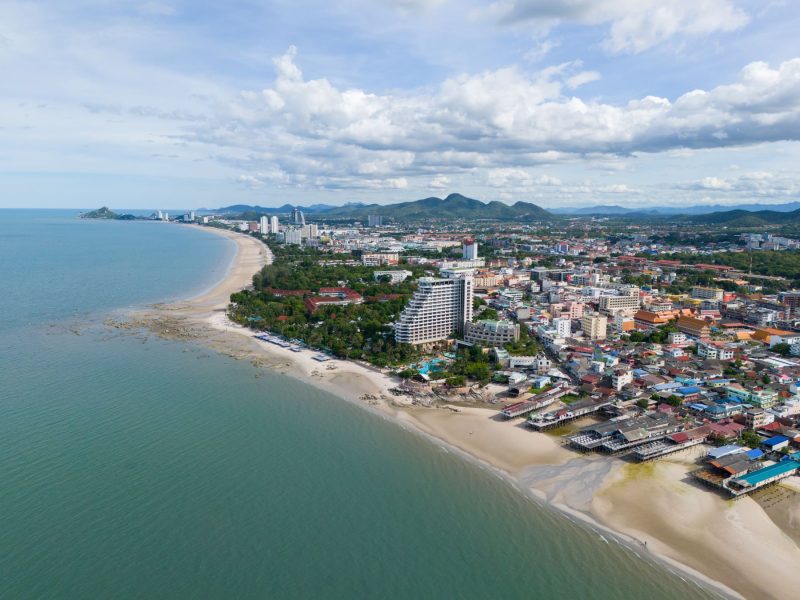 Aerial view of Hua Hin's coastline featuring sandy beaches, city buildings, and lush greenery under a partly cloudy sky.