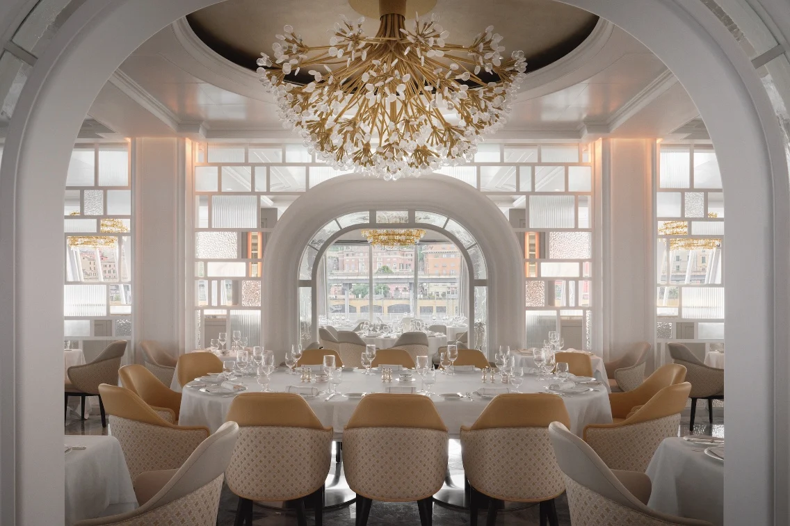 Elegant dining room with circular tables, plush chairs, and a decorative chandelier, featuring large windows for natural light.