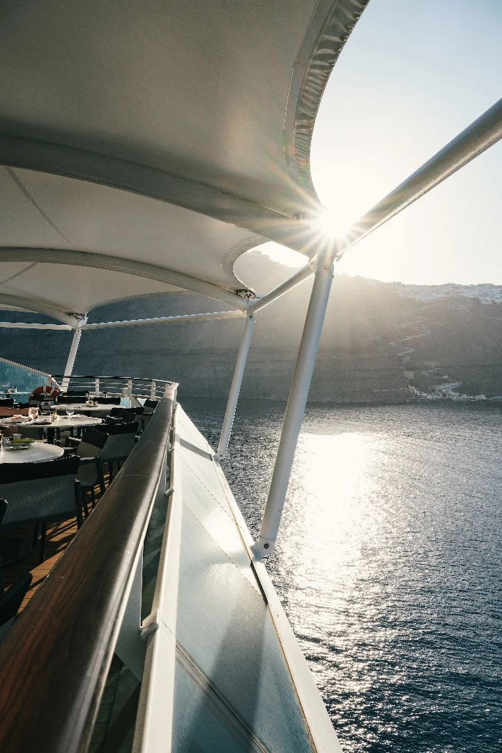 Sunlight glimmers on the water beside a terrace cafe deck with outdoor seating, offering a serene sunset view.