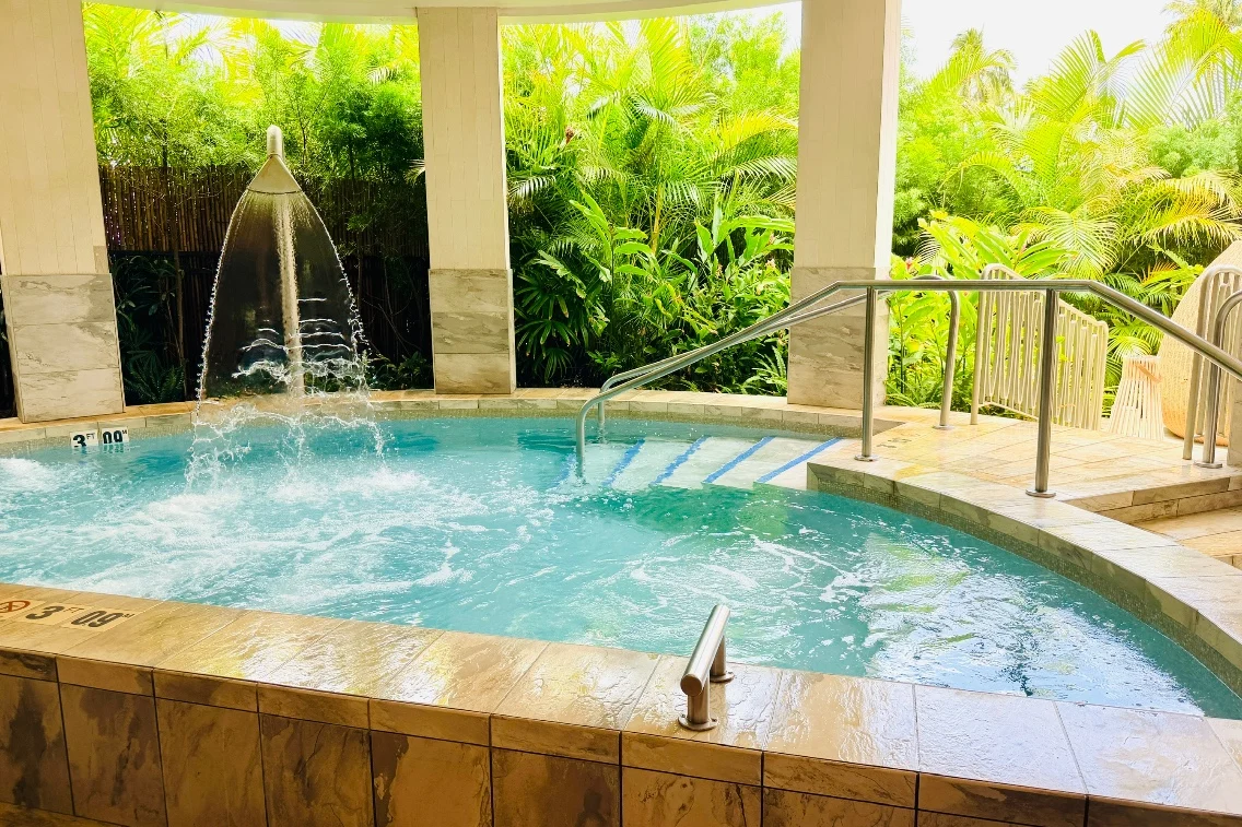Relaxing hot tub at Kilolani Spa surrounded by lush tropical plants, featuring a waterfall and accessibility railing.