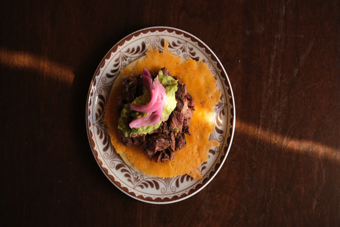 Beef taco topped with guacamole and pickled onions on a decorative plate.