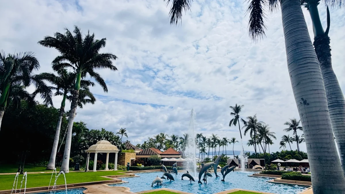 Pool area at Kilolani Spa, featuring dolphin sculptures, fountains, lush palm trees, and a cloudy sky at Grand Wailea Maui.