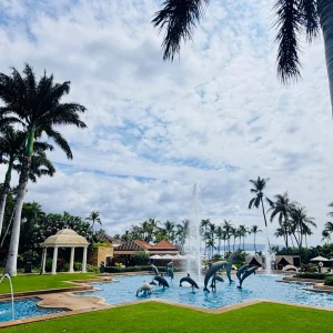 Pool area at Kilolani Spa, featuring dolphin sculptures, fountains, lush palm trees, and a cloudy sky at Grand Wailea Maui.