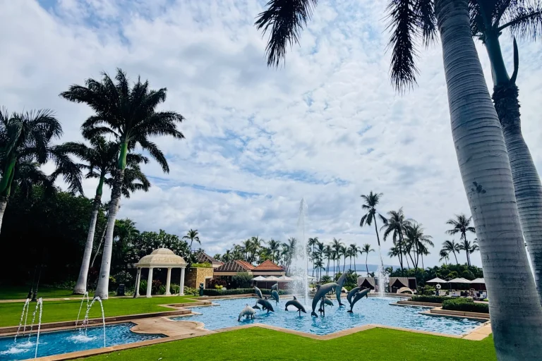 Pool area at Kilolani Spa, featuring dolphin sculptures, fountains, lush palm trees, and a cloudy sky at Grand Wailea Maui.