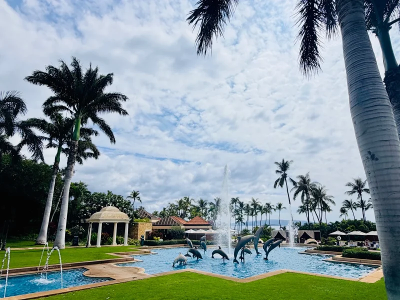 Pool area at Kilolani Spa, featuring dolphin sculptures, fountains, lush palm trees, and a cloudy sky at Grand Wailea Maui.