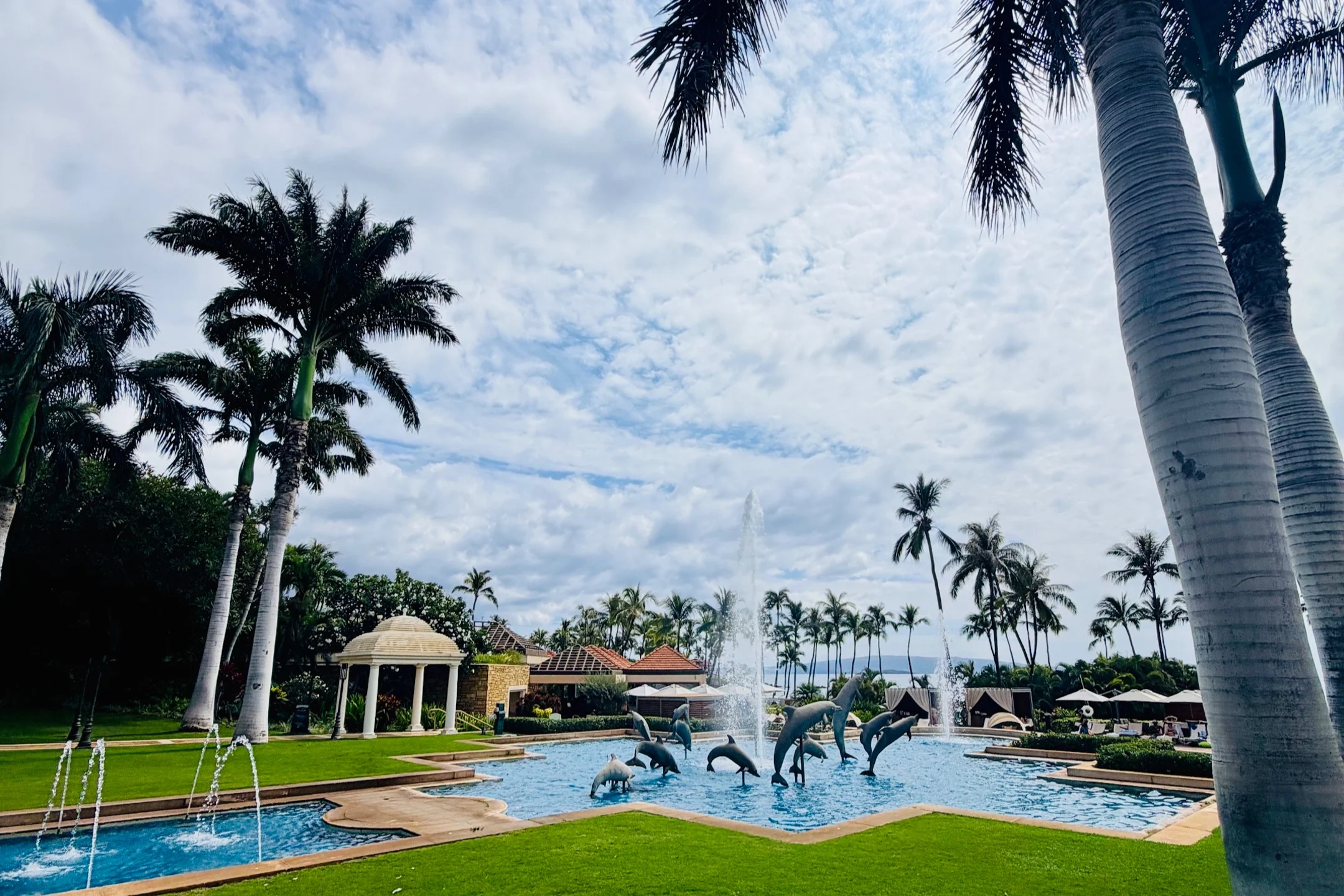 Pool area at Kilolani Spa, featuring dolphin sculptures, fountains, lush palm trees, and a cloudy sky at Grand Wailea Maui.
