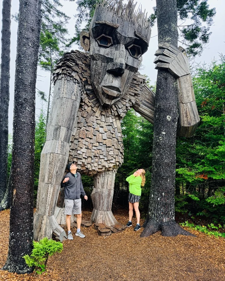 Giant wooden troll sculpture by Thomas Dambo with two people standing nearby in a forest setting.