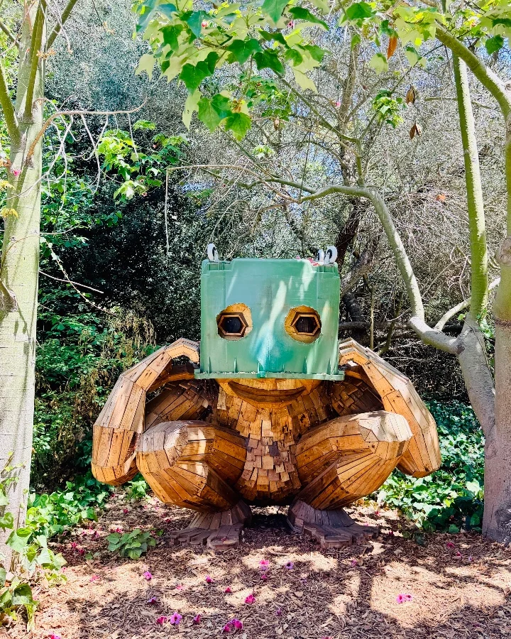 Wooden troll sculpture by Thomas Dambo, with a green box head and surrounded by greenery and flowers.