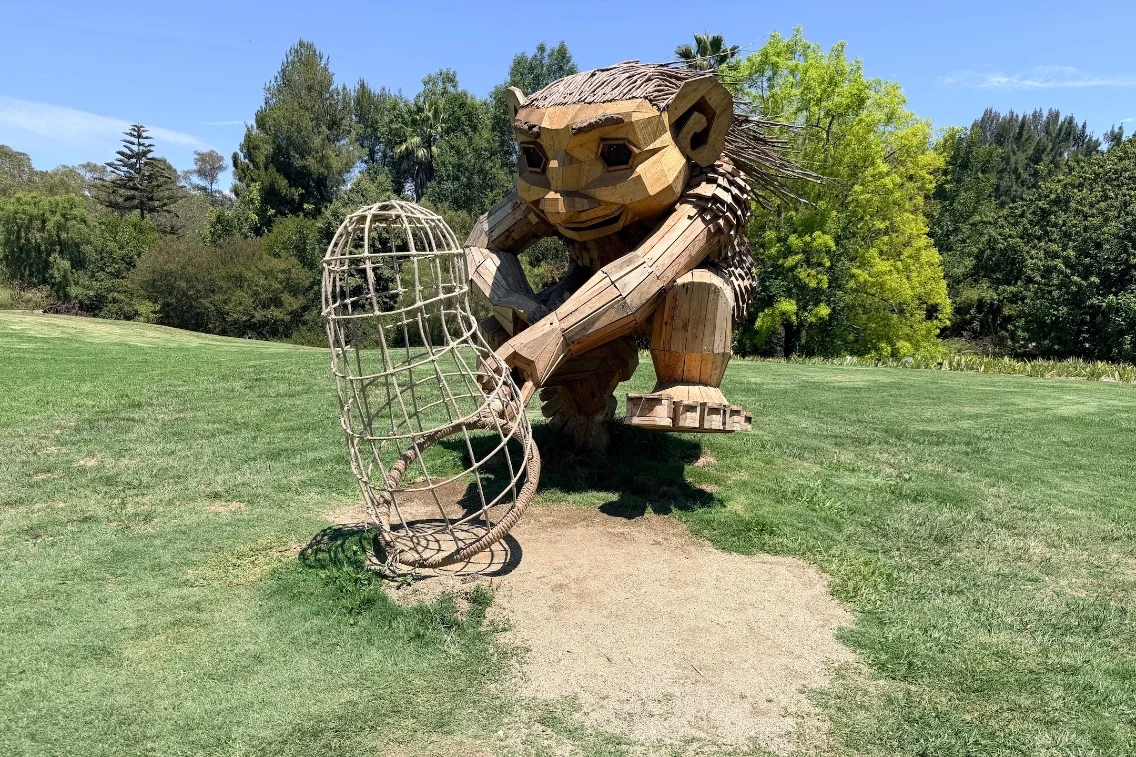 A large wooden troll sculpture crouches on the grass, holding a woven basket in its hands, surrounded by greenery.