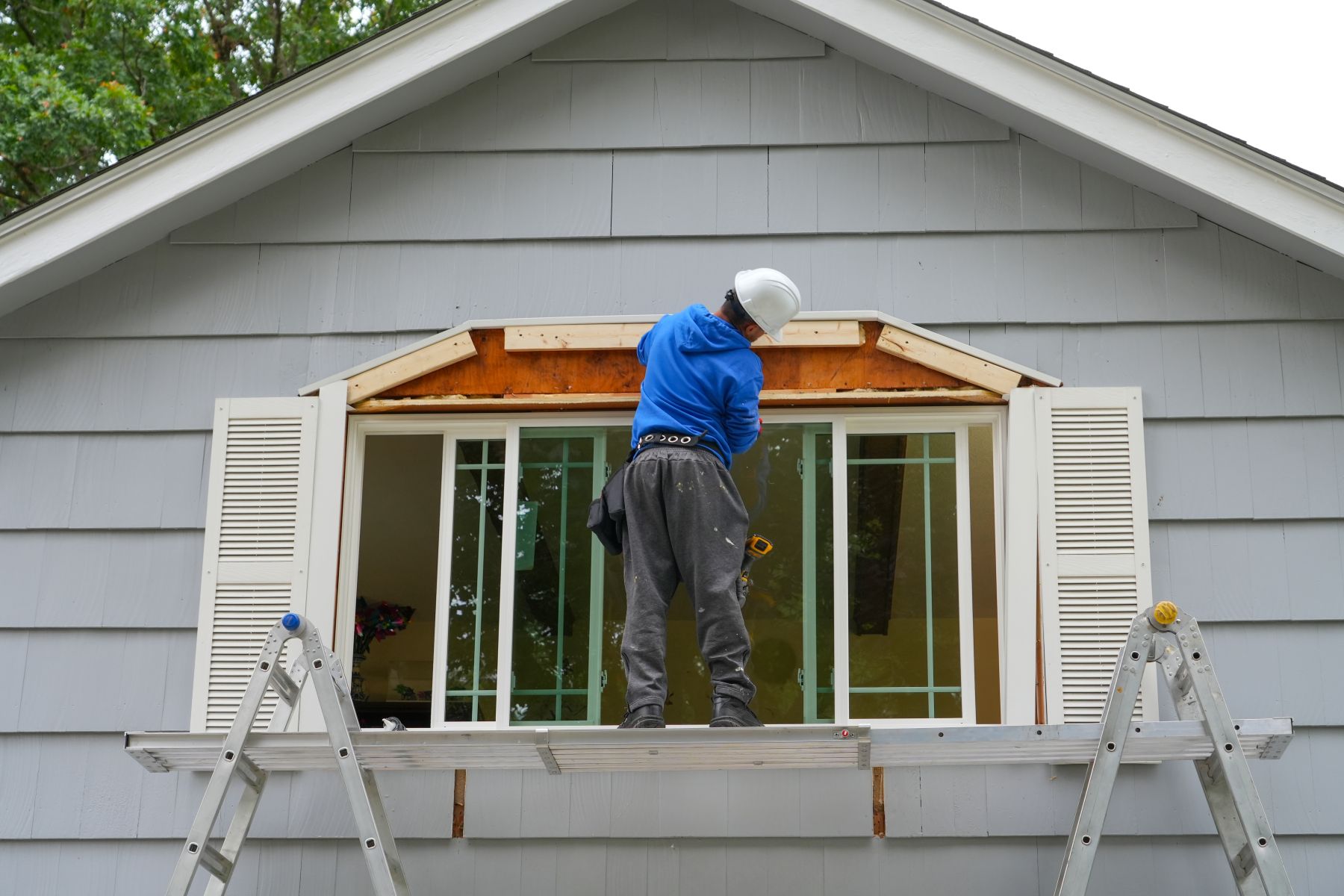 Worker installing a new window at a home, standing on a ladder with scaffolding and tools nearby.