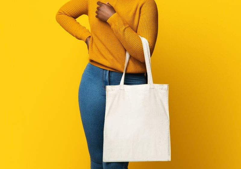 Young woman in a yellow sweater poses with a stylish white tote bag against a bright yellow background.