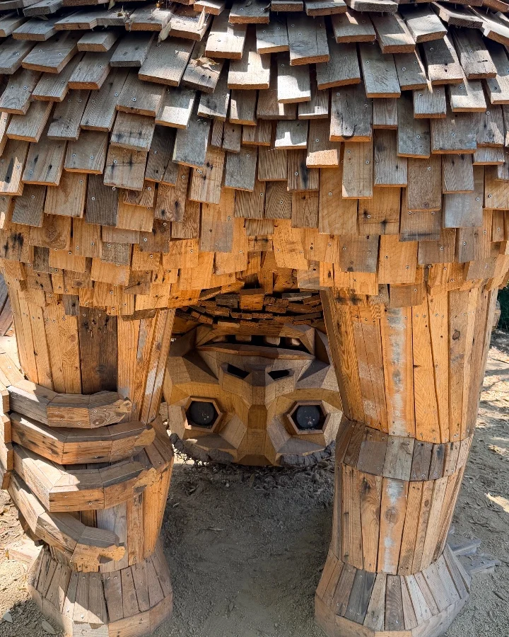 Wooden structure resembling a troll with a textured roof, large legs, and geometric shapes, showcasing recycled art.