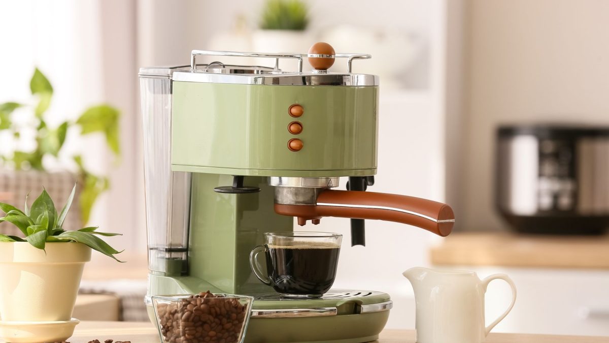 Green espresso machine brews coffee into a glass cup, surrounded by coffee beans and a small plant on a wooden countertop.