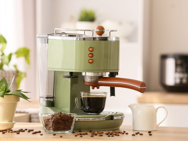 Green espresso machine brews coffee into a glass cup, surrounded by coffee beans and a small plant on a wooden countertop.