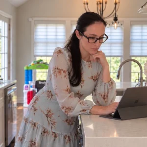 Kristin examines her tablet while standing at a bright kitchen counter adorned with flowers and kitchen items.