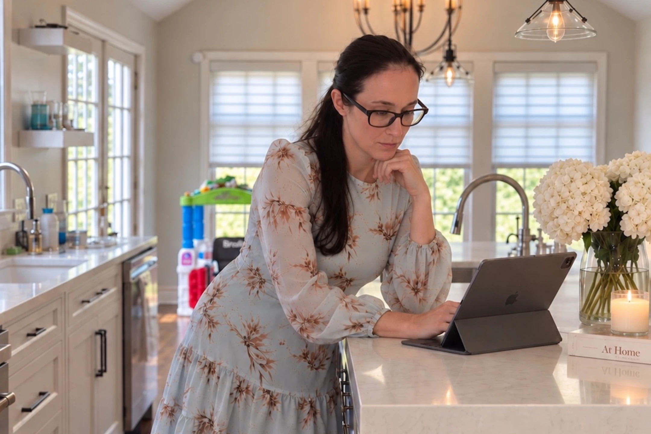 Kristin examines her tablet while standing at a bright kitchen counter adorned with flowers and kitchen items.
