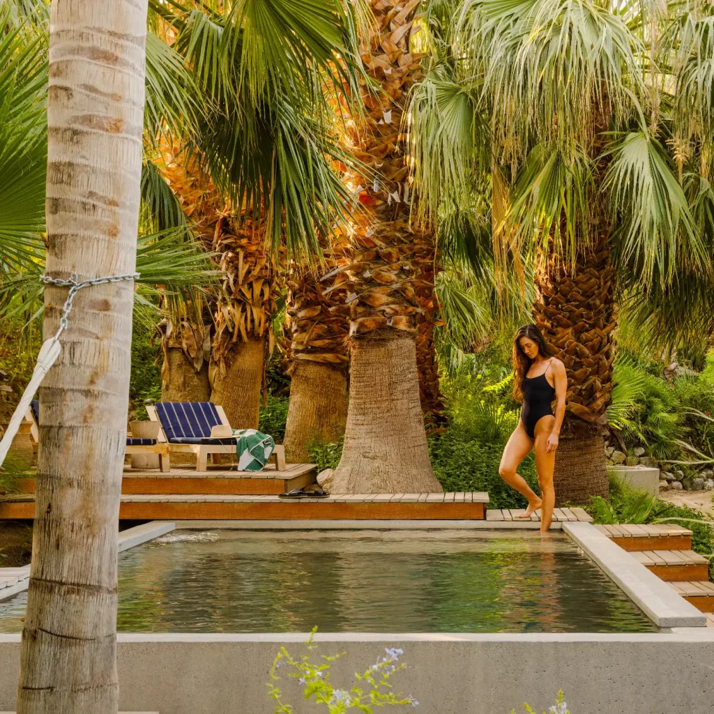 A woman walks near a serene pool surrounded by vibrant palm trees in a wellness retreat setting.
