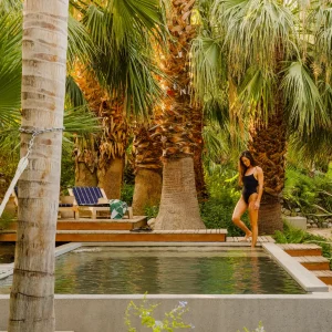 A woman walks near a serene pool surrounded by vibrant palm trees in a wellness retreat setting.