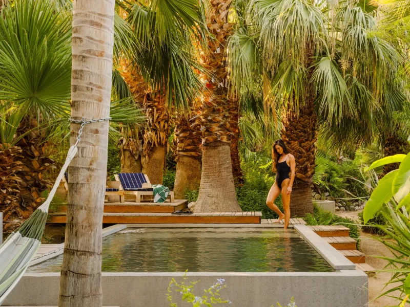 A woman walks near a serene pool surrounded by vibrant palm trees in a wellness retreat setting.