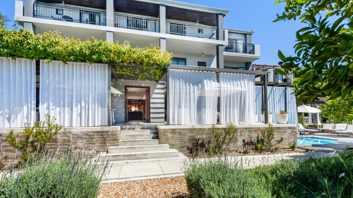 Modern building exterior with layered white curtains, steps leading to an entrance, and greenery surrounding a pool area.