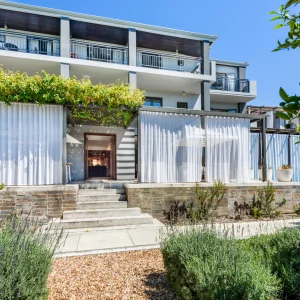 Modern building exterior with layered white curtains, steps leading to an entrance, and greenery surrounding a pool area.