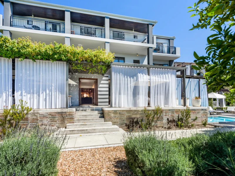 Modern building exterior with layered white curtains, steps leading to an entrance, and greenery surrounding a pool area.