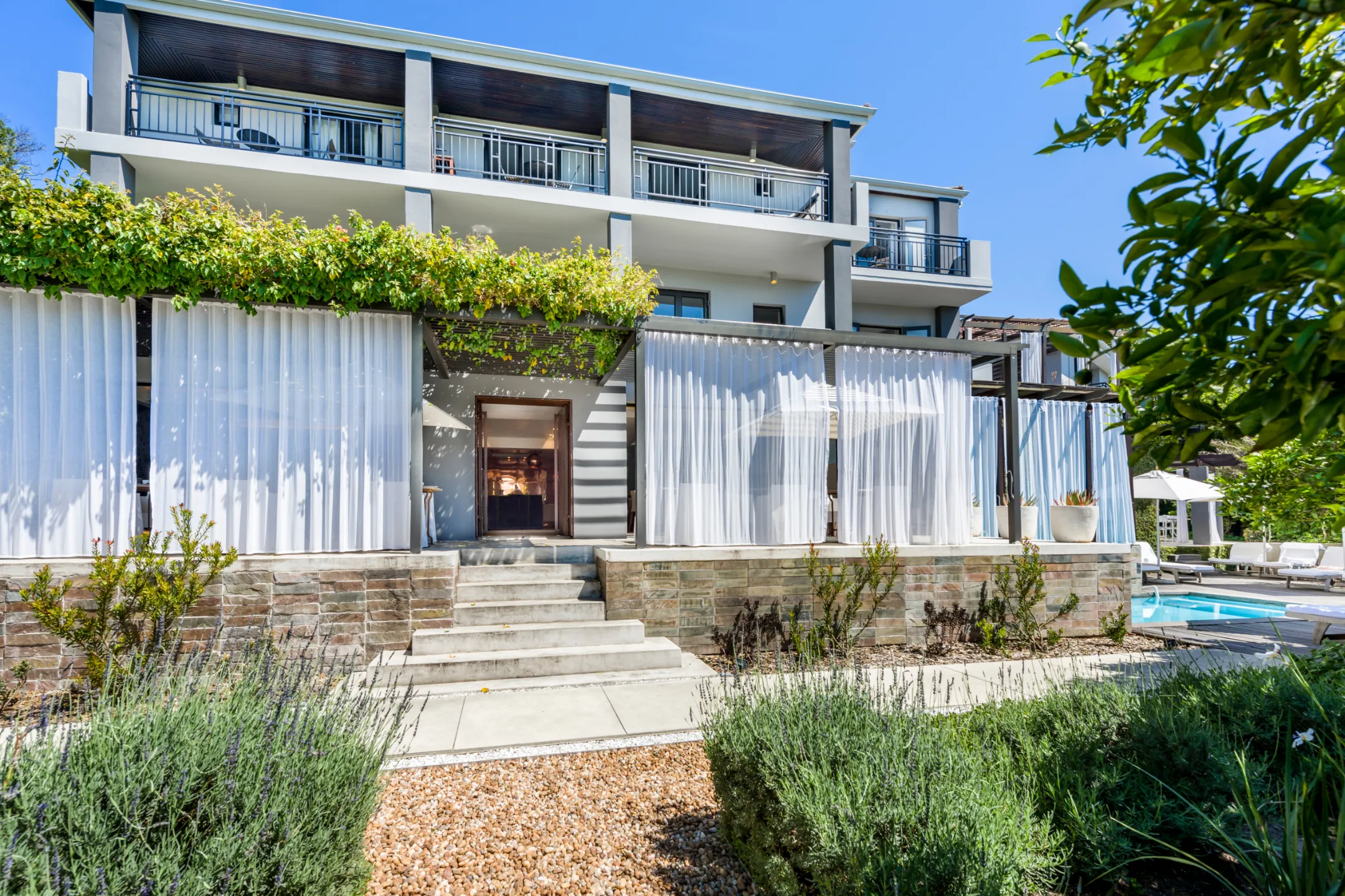 Modern building exterior with layered white curtains, steps leading to an entrance, and greenery surrounding a pool area.