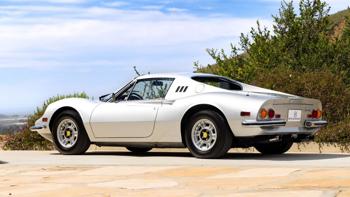 White Ferrari Dino 246 GTS parked outdoors, showcasing its sleek design and distinctive rear lights against a scenic backdrop.