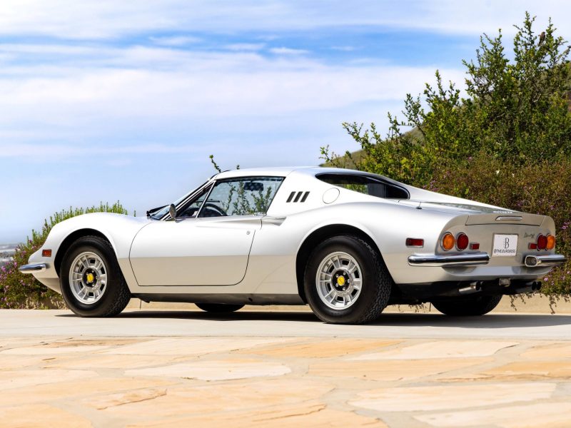 White Ferrari Dino 246 GTS parked outdoors, showcasing its sleek design and distinctive rear lights against a scenic backdrop.