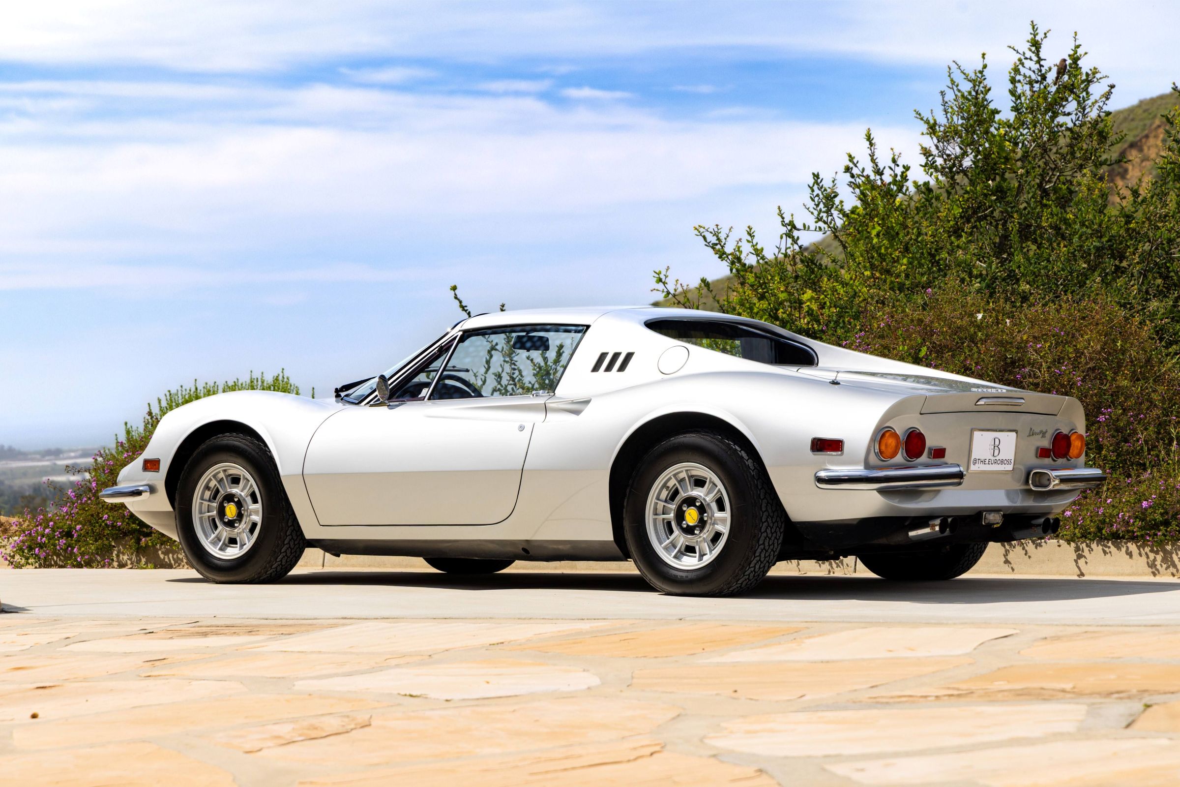 White Ferrari Dino 246 GTS parked outdoors, showcasing its sleek design and distinctive rear lights against a scenic backdrop.