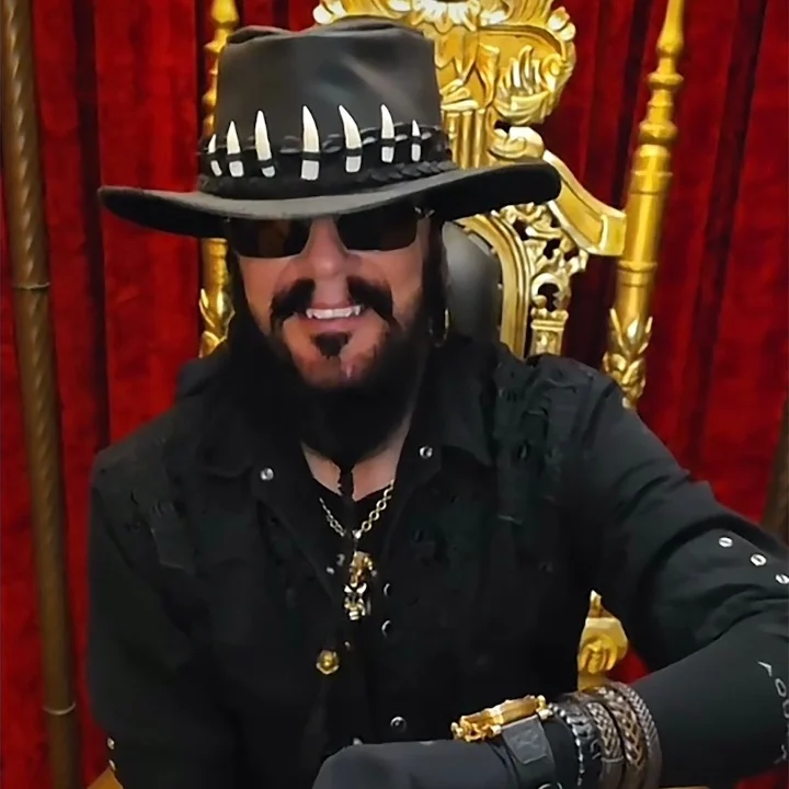 Man in a wide-brimmed hat and sunglasses smiles while seated on a throne, wearing a dark outfit with accessories and jewelry.