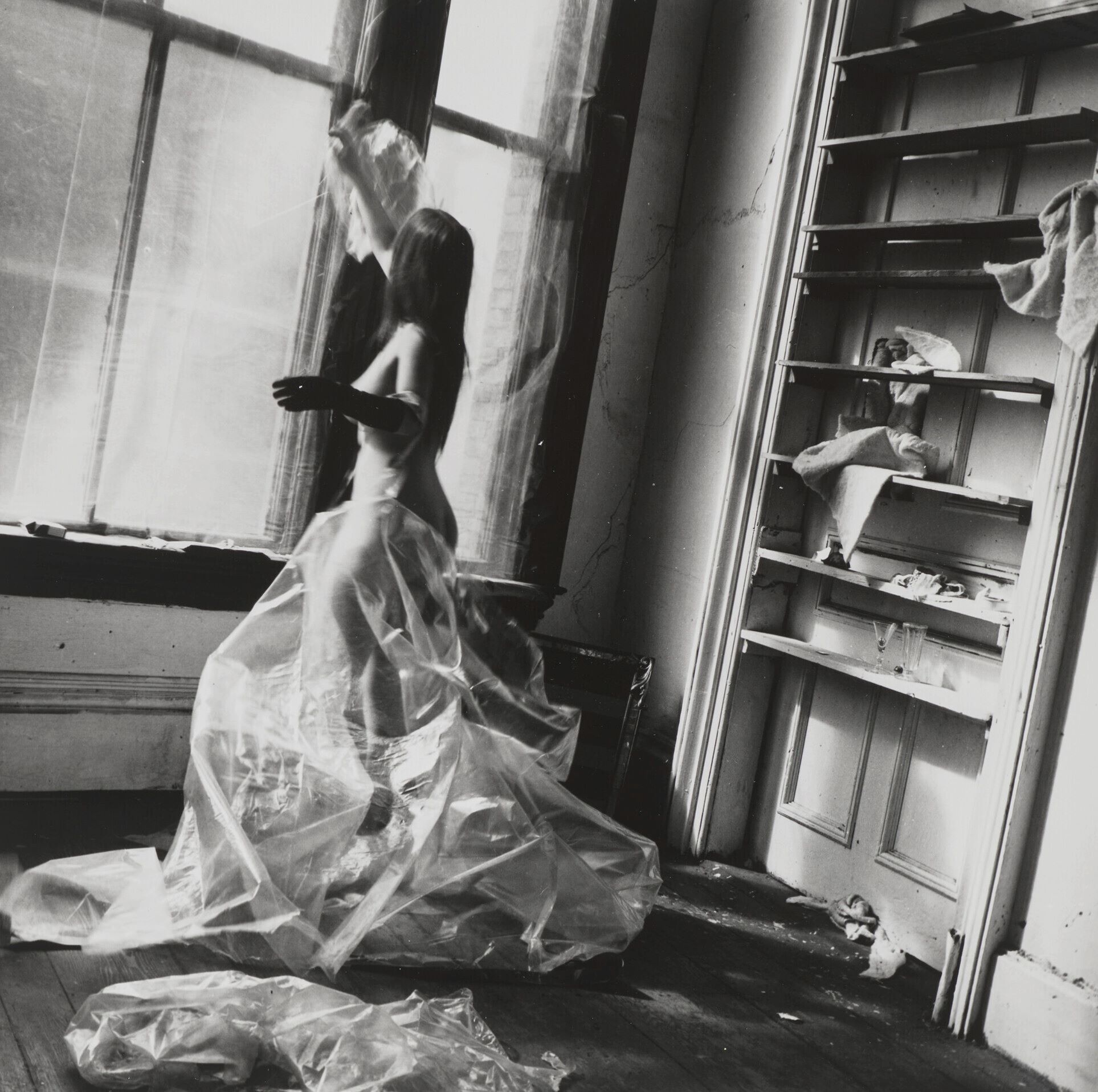 A figure draped in translucent plastic fabric stands by a window in a cluttered room, creating a striking contrast.