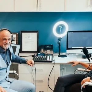 Two hosts with headphones sit at a desk with a microphone and production equipment, preparing to record a podcast.