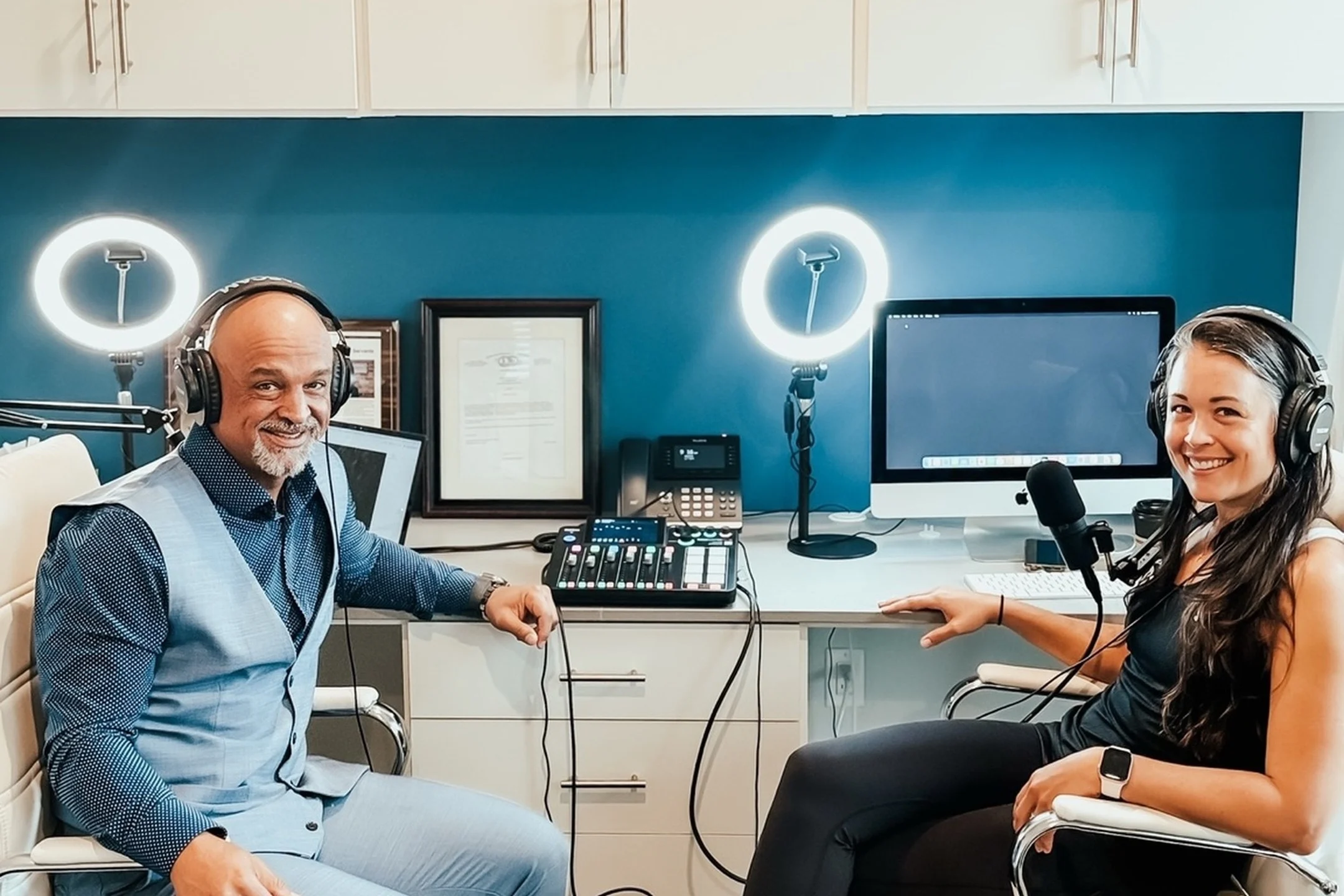Two hosts with headphones sit at a desk with a microphone and production equipment, preparing to record a podcast.