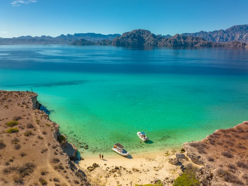 Clear turquoise waters surround a sandy beach, with two boats anchored and rocky cliffs in the distance under a bright blue sky.