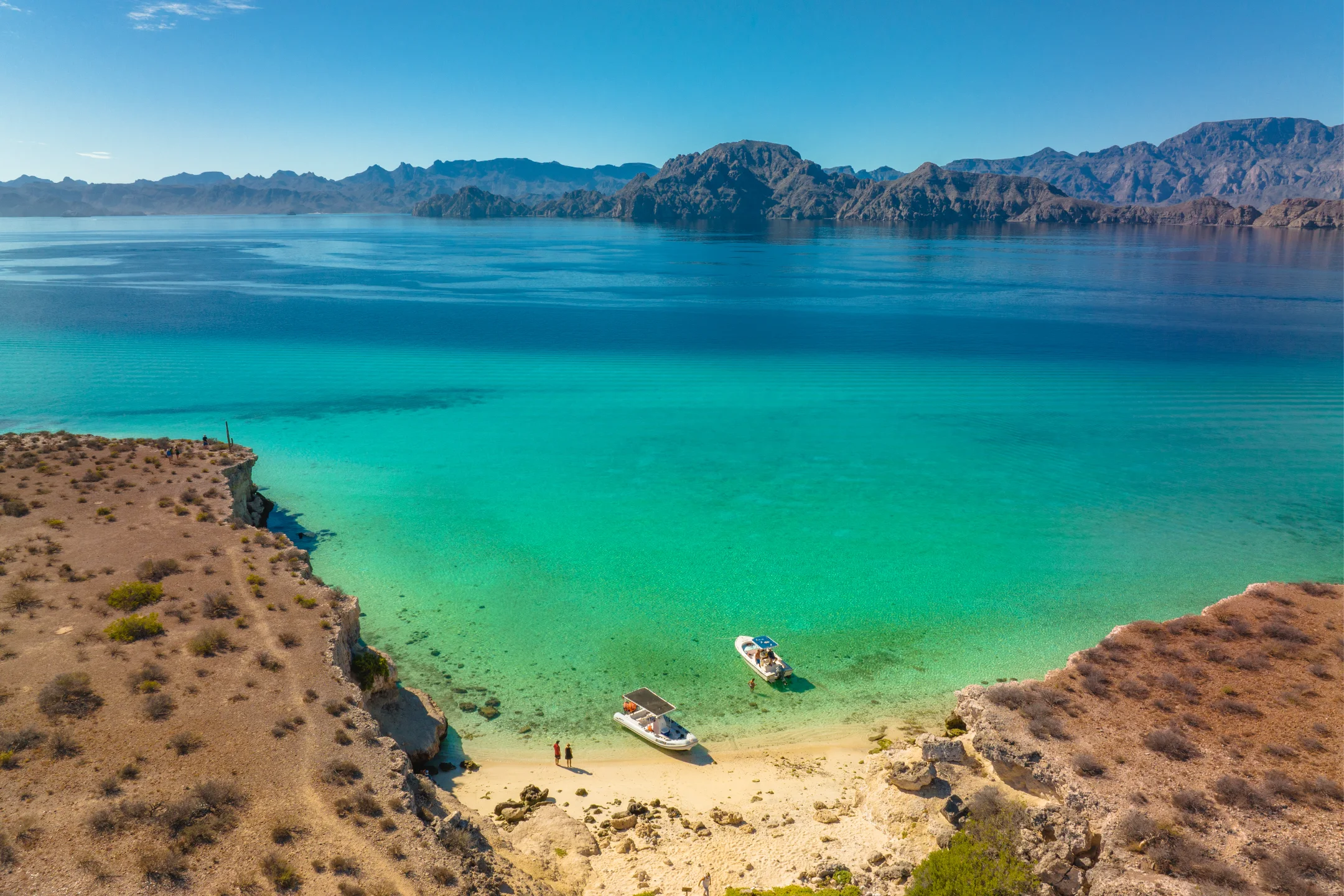 Clear turquoise waters surround a sandy beach, with two boats anchored and rocky cliffs in the distance under a bright blue sky.