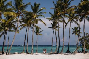 Two women walk along a sandy beach lined with palm trees, overlooking the clear blue ocean at St. Regis Cap Cana Resort.