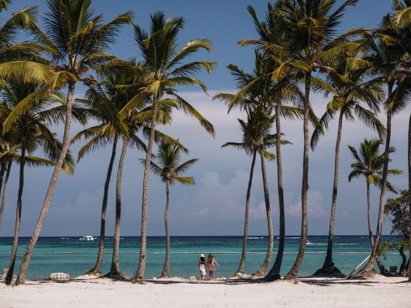 Two women walk along a sandy beach lined with palm trees, overlooking the clear blue ocean at St. Regis Cap Cana Resort.